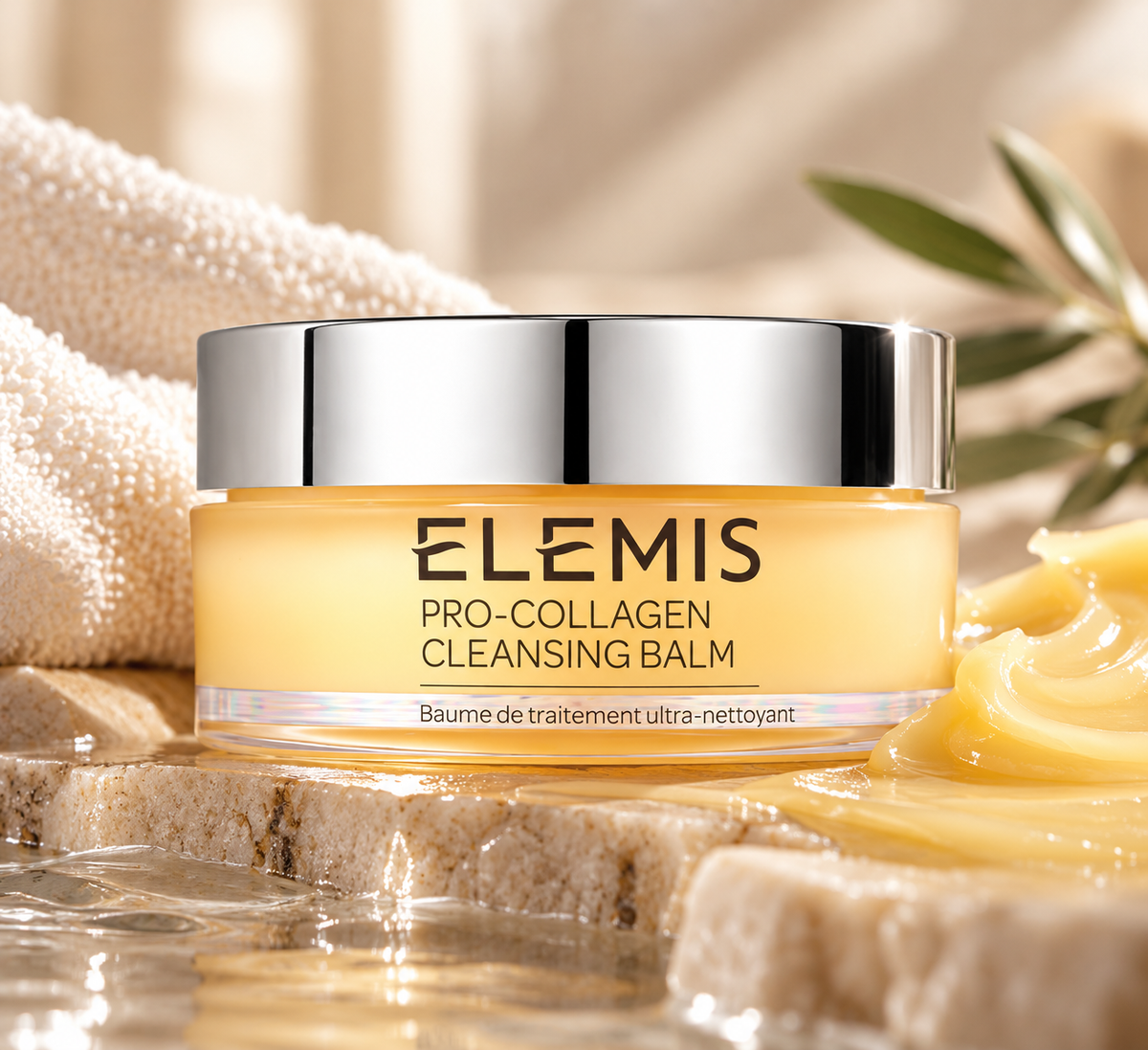 ELEMIS Pro-Collagen Cleansing Balm luxury facial cleanser in a golden jar with a silver lid, elegantly displayed on natural stone beside soft white towels, botanical leaves and a rich balm texture. Nourishing cleansing balm designed to melt away makeup, impurities and daily buildup while leaving skin soft and hydrated.