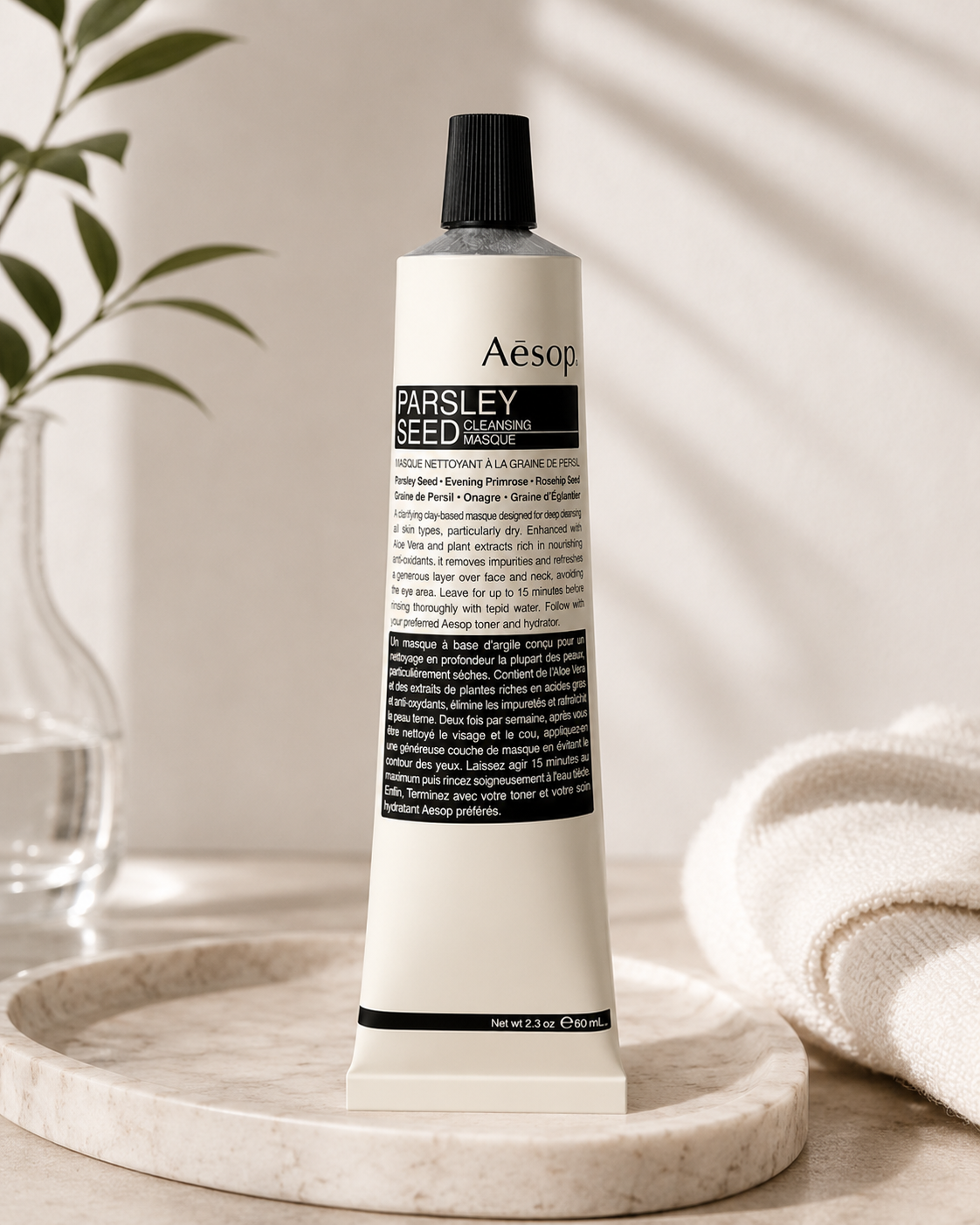 Aesop Parsley Seed Cleansing Masque displayed upright on a light stone tray in a minimalist spa-inspired bathroom with soft natural light, a glass vase with greenery, and folded white towels. The calming setting highlights the antioxidant-rich clay facial mask as part of a luxurious skincare ritual, emphasizing deep cleansing, skin purification, botanical ingredients, and Aesop's refined apothecary aesthetic.